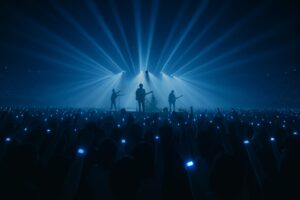 巨大な会場でライブバンドのシルエットが青い光に包まれ、観客の腕には青く光るリストバンドが一面に広がっている様子。